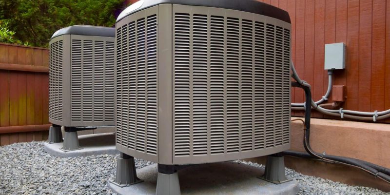 2 HVAC units installed near the outside of a home. The area is surrounded by rocks and wood paneling